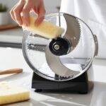 Food processor blade being cleaned with a soft sponge and warm water, with soap bubbles and water droplets visible on the shiny stainless-steel blade. The kitchen counter features dish soap, a small brush, and a soft sponge, highlighting the importance of maintaining kitchen tools.